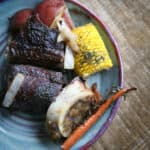 Mexican beef short ribs recipe by Yvette Marquez-Sharpnack of Muy Bueno served with potatoes, carrots, corn, and marrow bone on a rustic plate