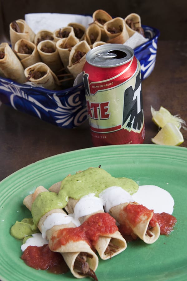 three falutas filled with shredded beef on a green plate drizzled with salsa Crema avocado salsa banderas taquitos with a Tecate in the background