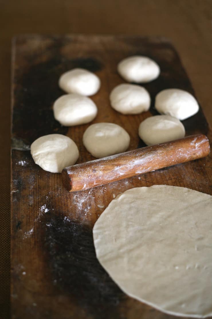 One tortilla, a small wooden rolling pin and eight testales bolts balls of dough on a wooden board