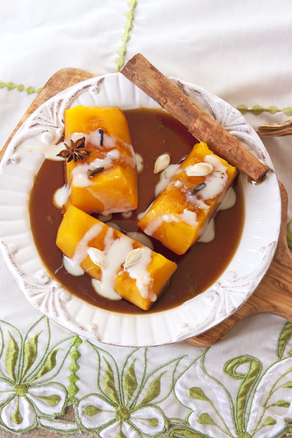 Calabaza en Tacha a.k.a. Candied Pumpkin with pilloncillo syrup and drizzled with sweetened condensed milk on a white plate atop a wooden cutting board on a sage green and white embroidered tea towel