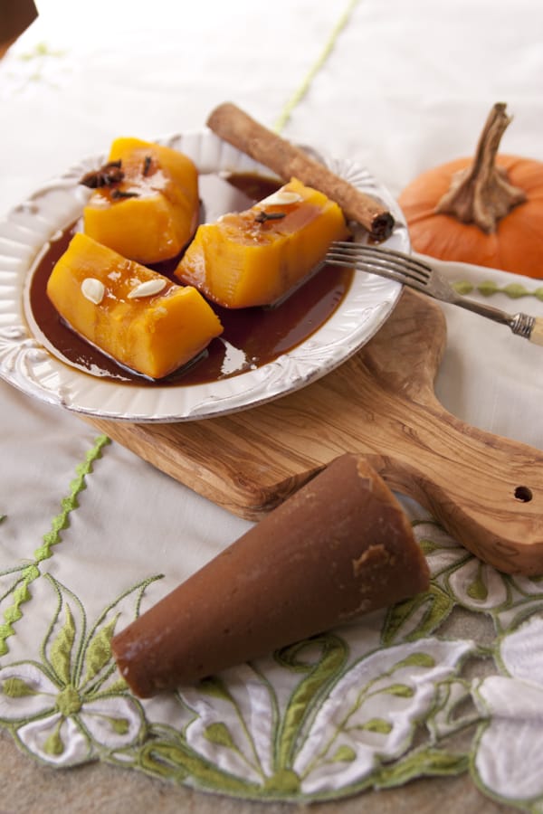 45 degree angle shot of mexican candied pumpkin wedges on a white plate with a puddle of brown syrup and drizzled with sweetened condensed milk on a white plate on a wooden cutting board with a pumpkin and a cinnamon stick