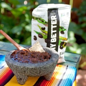 Chile pasilla Oaxaqueño salsa in a stone molcajete with a wooden spoon, served with tortilla chips on a colorful Mexican textile.