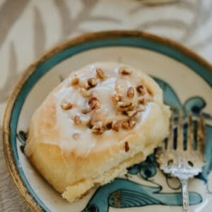 Close-up of a glazed dulce de leche cinnamon roll topped with chopped pecans on a decorative ceramic plate.