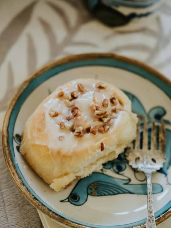 Close-up of a glazed dulce de leche cinnamon roll topped with chopped pecans on a decorative ceramic plate.