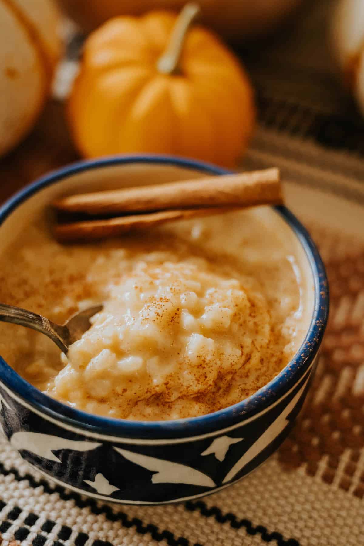 Close-up of Mexican Pumpkin Rice Pudding in a clay bowl with a pumpkin behind it, showing creamy texture and cinnamon garnish.