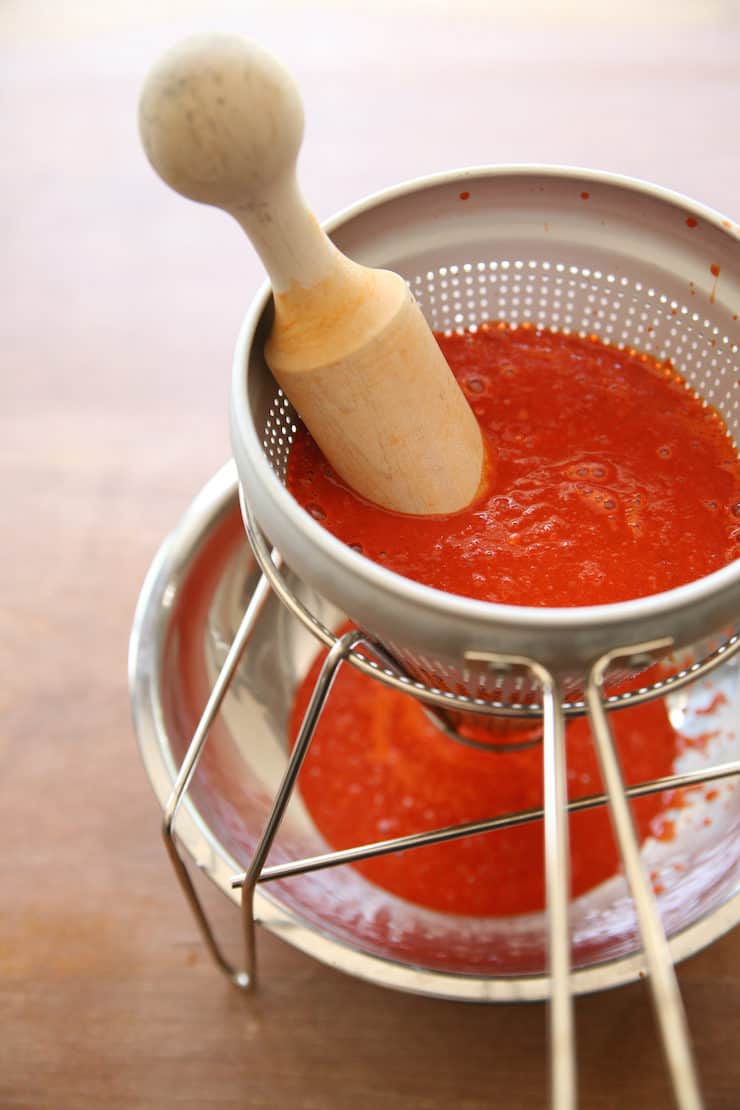 homemade enchilada sauce in a conical sieve with a pestle resting in a conical sieve with homemade red chile sauce straining through it into a silver bowl