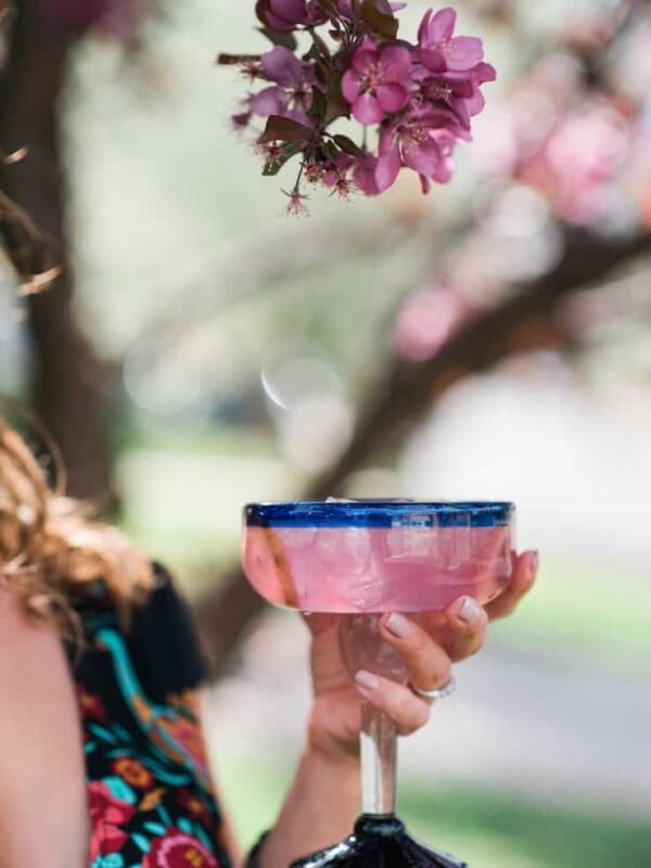 Woman holding a pink prickly pear margarita in a colorful Mexican glass beneath blooming pink flowers, inspired by the desert flavors of El Paso, Texas.