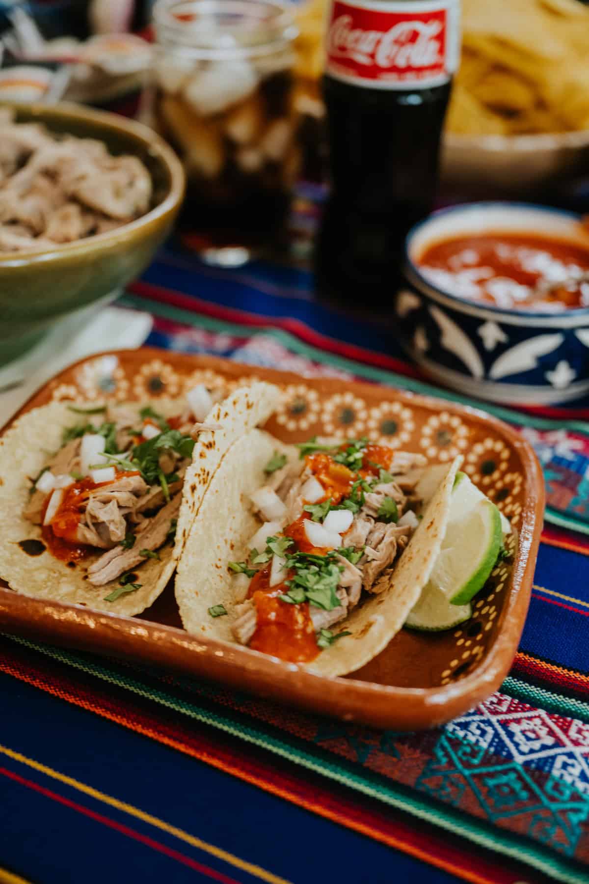 Two carnitas tacos topped with onion, cilantro, and salsa served on a Talavera-style plate next to a bottle of Mexican Coca-Cola.