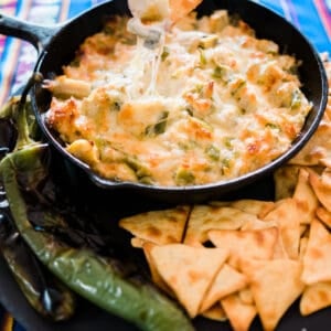 hand dipping a pita chip into cheesy Green Chile Artichoke Dip in a black skillet surrounded by chips.