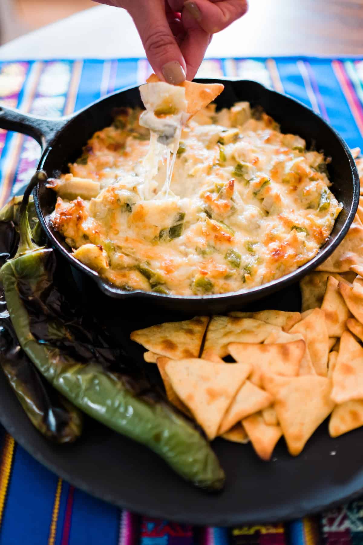 hand dipping a pita chip into cheesy Green Chile Artichoke Dip in a black skillet surrounded by chips.