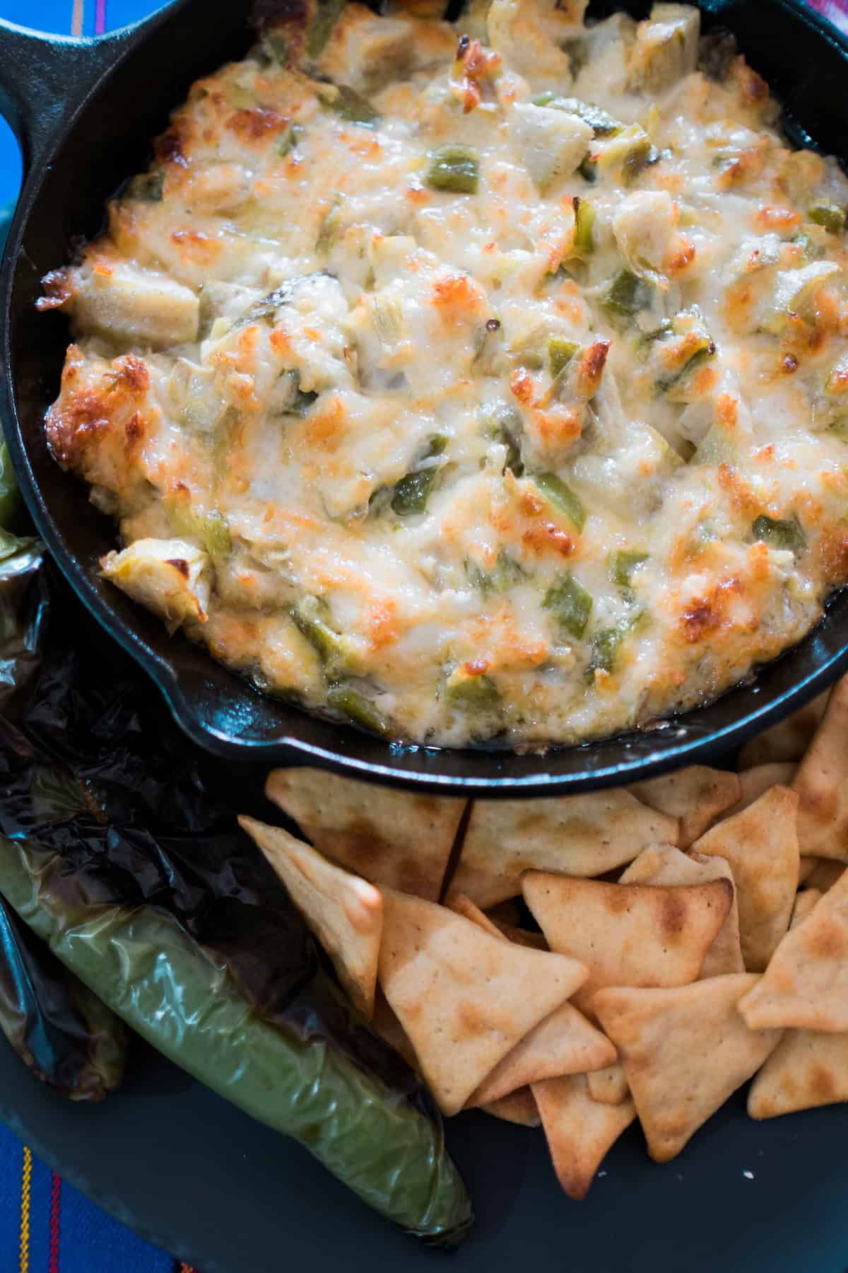 Cheesy Green Chile Artichoke Dip in a black skillet surrounded by chips and roasted green chile peppers.