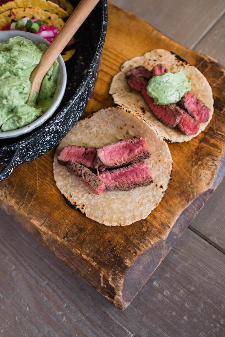 sliced bison steak on corn tortillas on a cutting board and topped with avocado crema