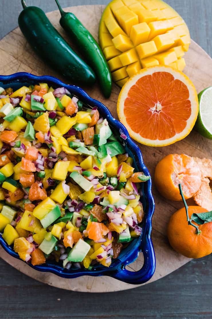 overhead shot of a blue scalloped bowl of orange, avocado and mango salsa on wooden cutting board next to fresh jalapeños, mango, cara cara orange, lime and tangerine