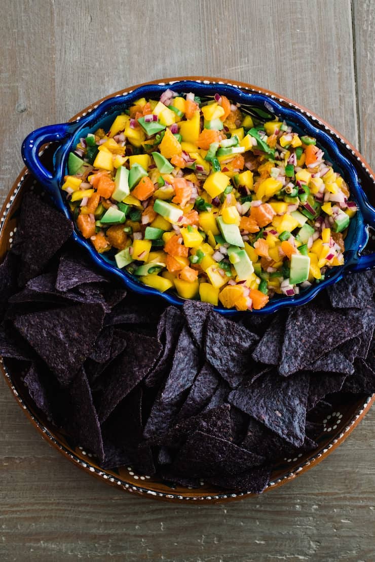 blue serving bowl of orange, avocado and mango salsa on a serving tray filled with blue corn tortilla chips