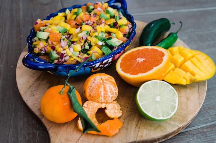 blue bowl of orange, avocado and mango salsa on a cutting board with the raw ingredients to make it