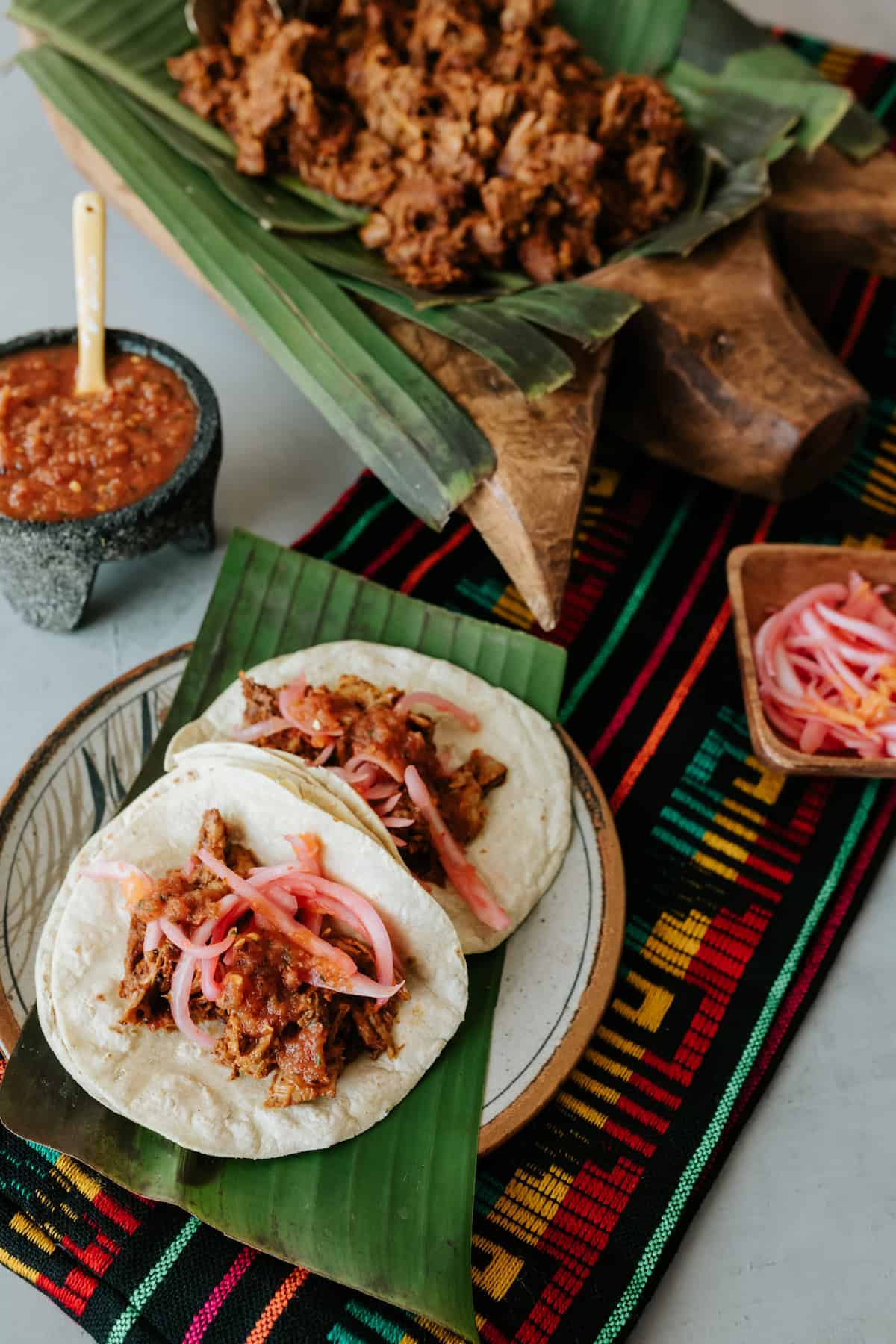 Two Cochinita Pibil Tacos topped with pickled red onions and salsa, served on a plate with banana leaf over a colorful Mexican textile.