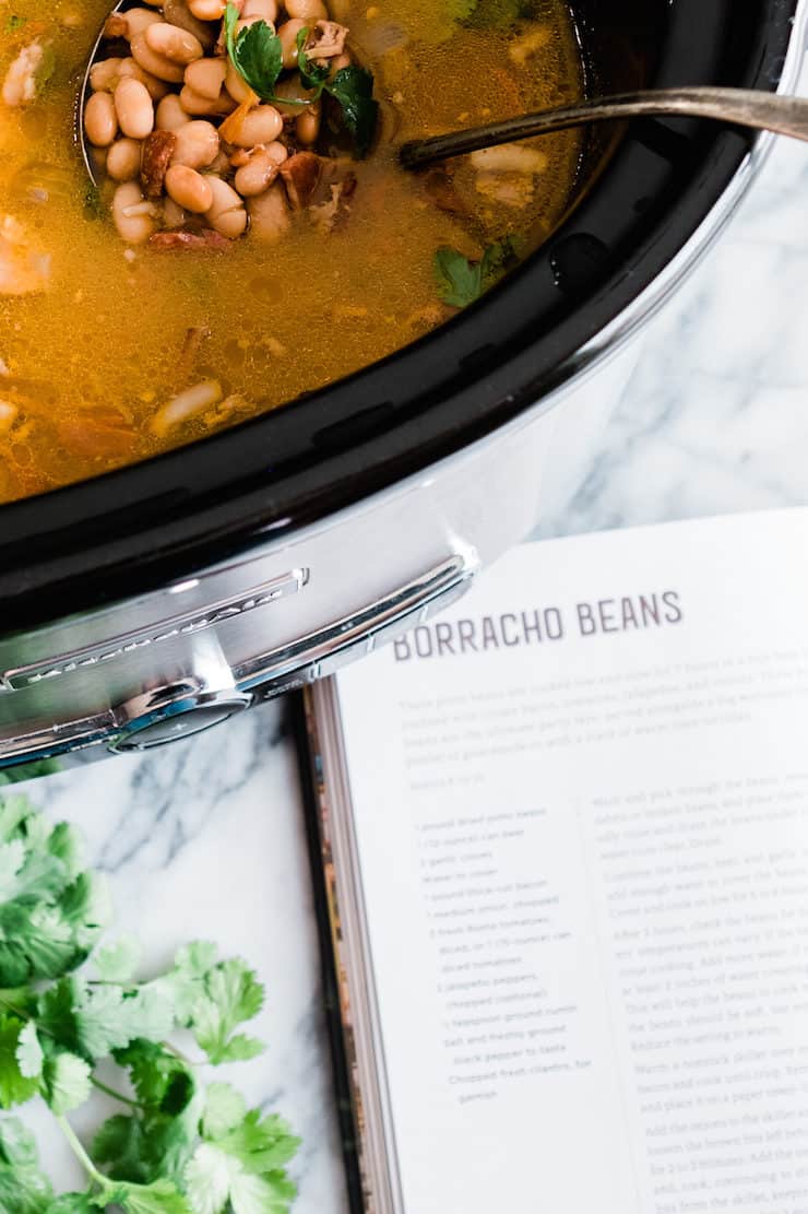 crockpot filled with borracho beans next to cookbook turned to recipe page