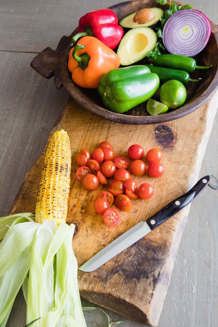 wooden bowl of veggies on a cutting board with an ear of roasted corn and some halved cherry tomatoes