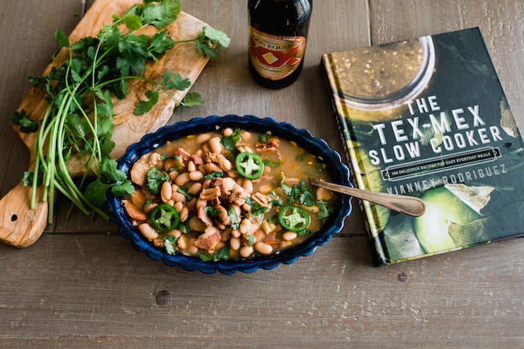blue scalloped serving dish of borracho beans next to "Tex Mex Slow Cooker" cookbook, bottle of beer and a wooden cutting board of fresh cilantro