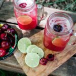 Mason jar mugs filled with Cherry Limeade Margaritas on a cutting board with line slices and bowl of cherries