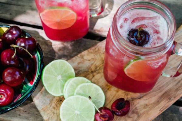 Mason jar mugs filled with Cherry Limeade Margaritas on a cutting board with line slices and bowl of cherries