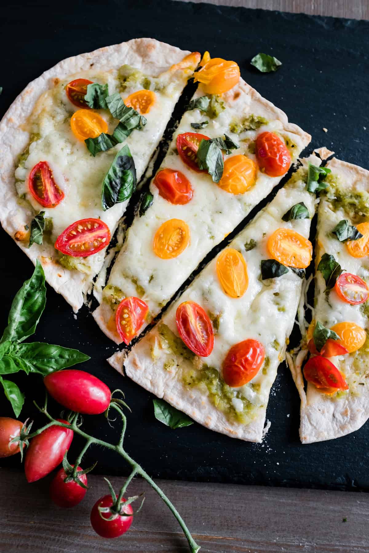 Overhead view of a sliced homemade Mexican pizza topped with melted cheese, yellow and red cherry tomatoes, fresh basil, and salsa verde, served on a black slate with a vine of fresh tomatoes and basil leaves on the side.