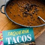 Frijoles Adobados ready in a big dish with Taqueria Tacos cookbook right next to it on a big wooden board