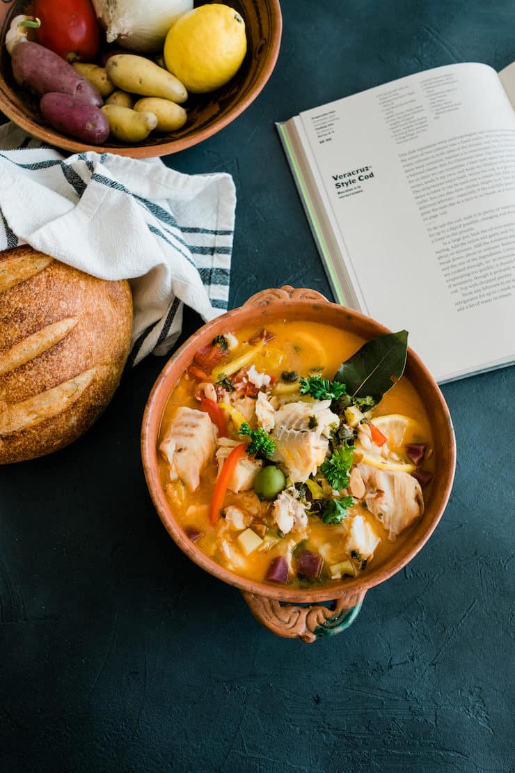 Veracruz-Style seafood stew looking absolutely delicious with bread on the left and vegetables in a big bowl next to the open cookbook