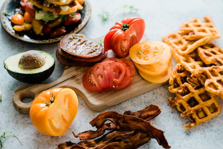wooden cutting board with sliced heirloom tomatoes, half an avocado, crispy bacon, and cornmeal waffles. An assembled BLT Waffle is blurred in the back of the image.