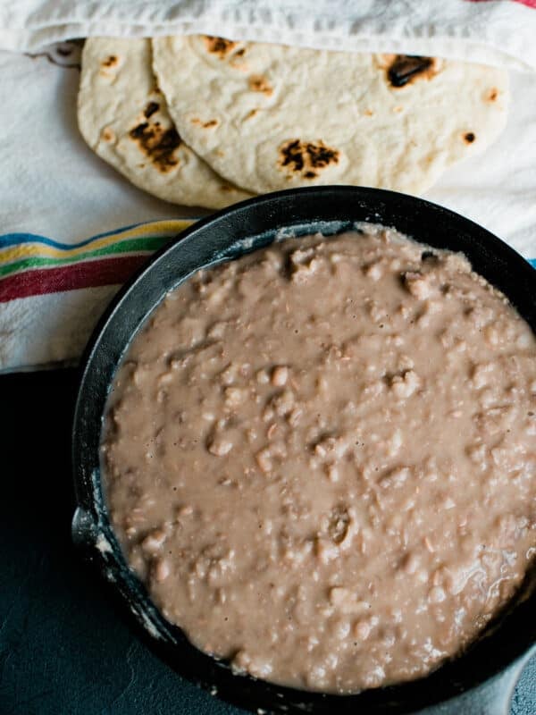 Homemade refried beans in a cast-iron skillet next to freshly made flour tortillas wrapped in a striped kitchen towel.