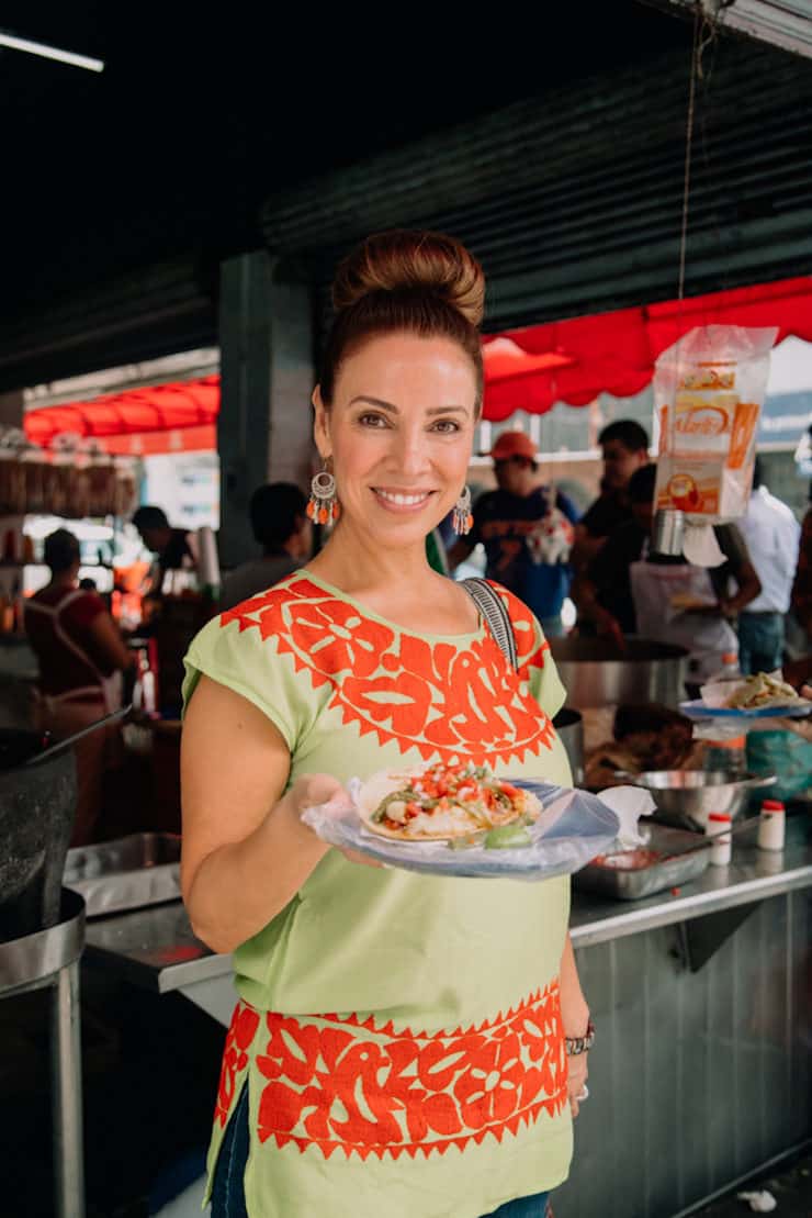 food and travel blogger Latina Yvette Marquez in Mexico City holding a plate of tacos wearing a colorful embroidered green and orange Mexican blouse