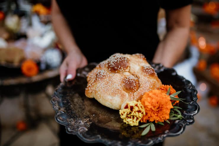 latina holding a black plate with two elements of dia de los muertos: special bread an marigolds for a day of the dead celebration