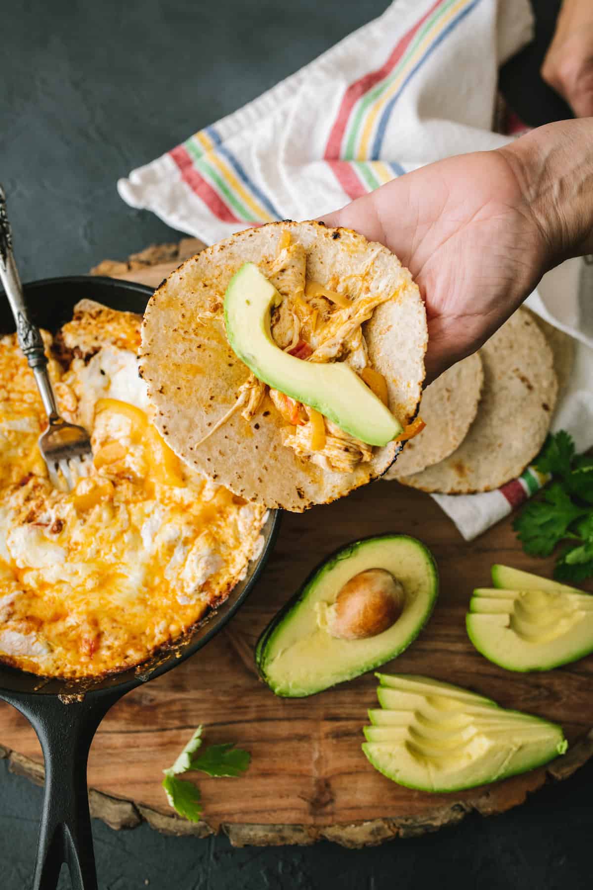 Hand holding a corn tortilla filled with melted queso fundido, sautéed bell peppers, shredded chorizo mixture, and a fresh avocado slice, with a cast-iron skillet of bubbling cheese dip in the background.