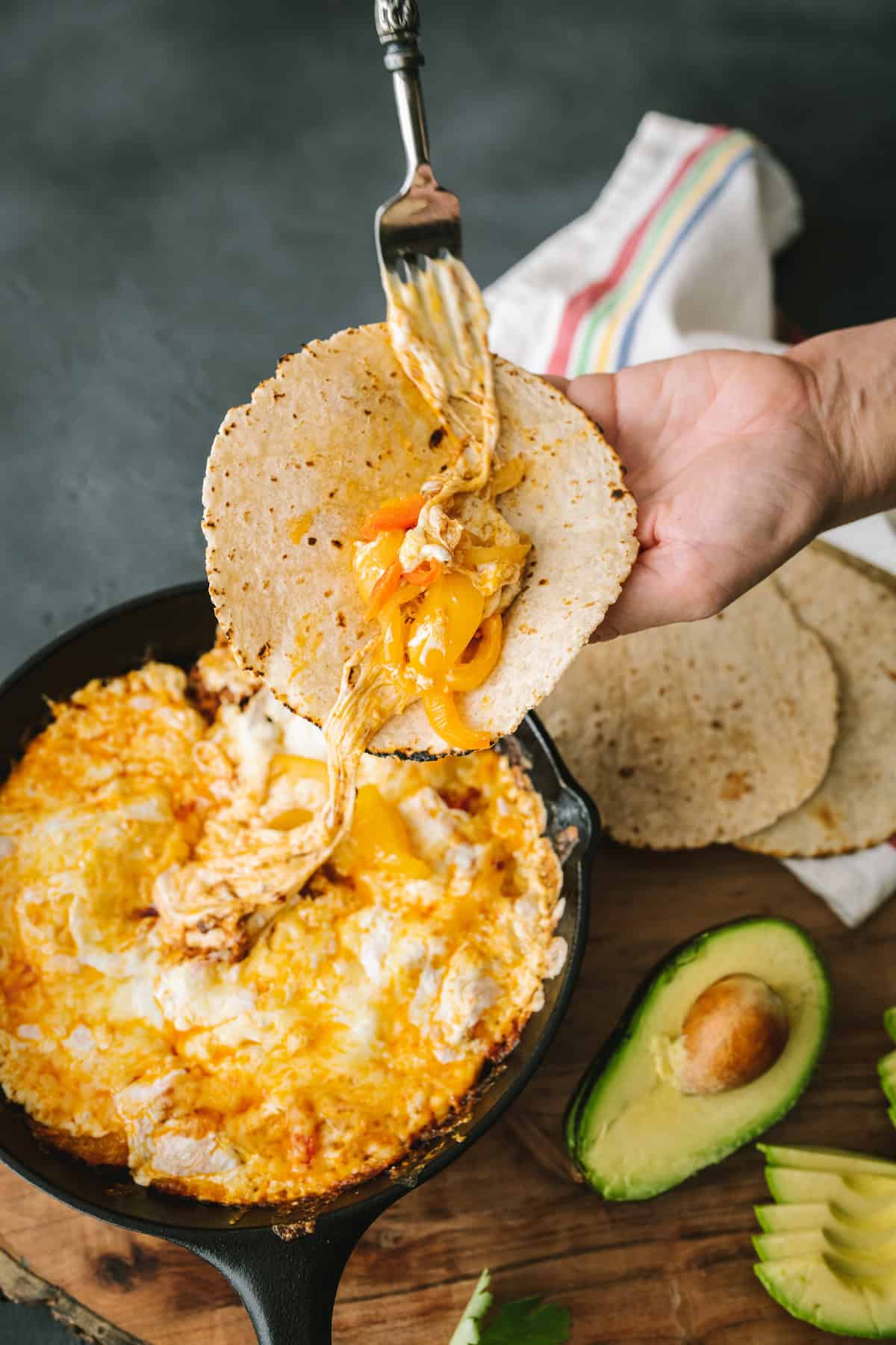 Fork placing gooey chorizo cheese mixture onto a warm corn tortilla over a skillet of hot, melted queso fundido, with colorful bell peppers and cheese stretching from the skillet.