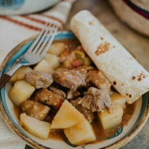 Close-up of tender beef and potatoes in chile verde sauce with a rolled corn tortilla and fork beside the plate.