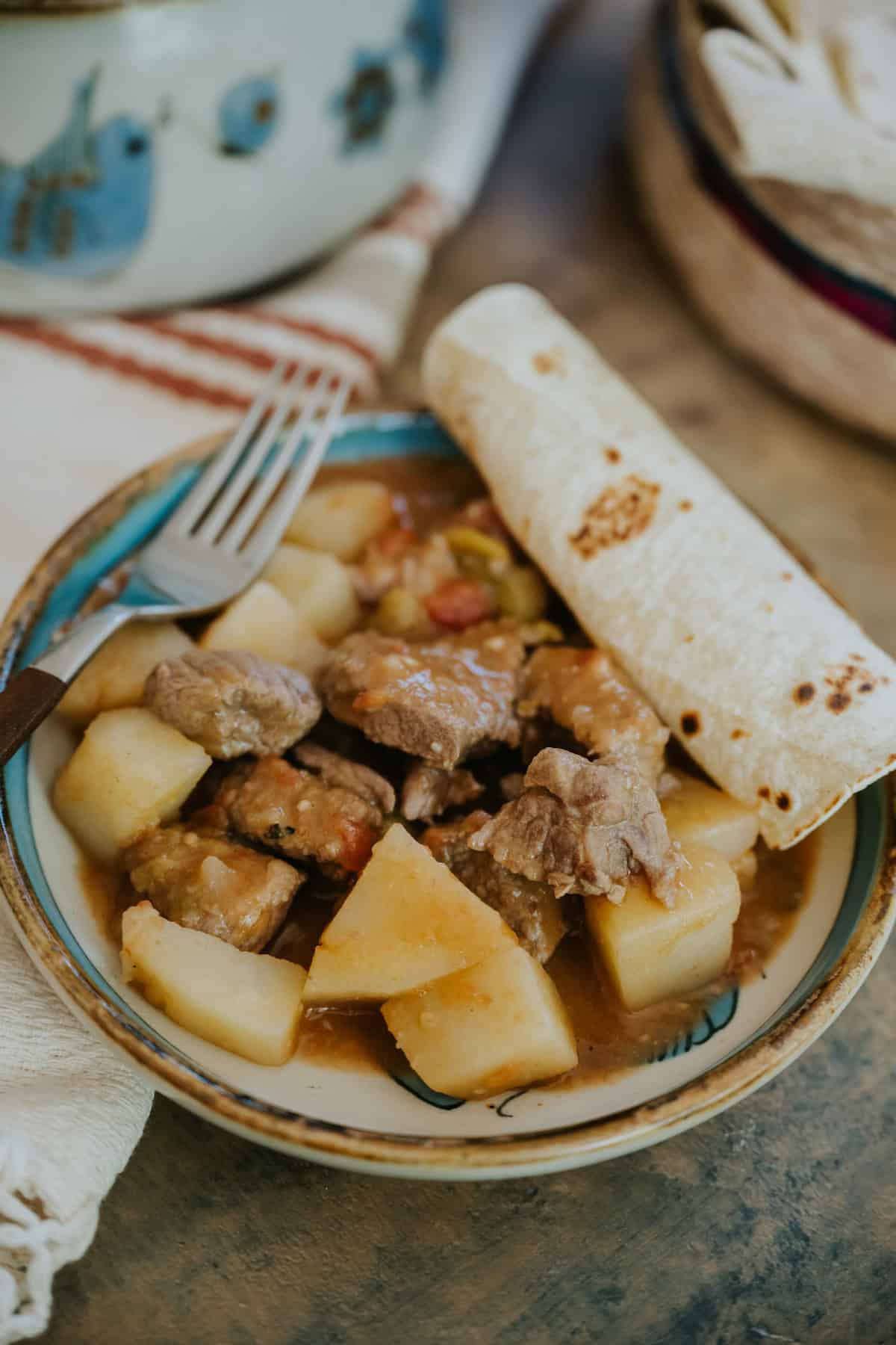 Close-up of tender beef and potatoes in chile verde sauce with a rolled corn tortilla and fork beside the plate.