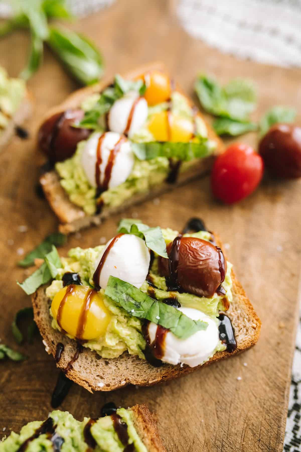 Overhead view of Caprese Avocado Toast slices on a wooden board, topped with colorful tomatoes, mozzarella, basil, and balsamic glaze.