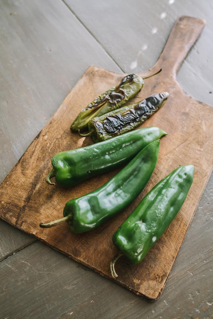 two blackened, roasted peppers on a cutting board with three fresh peppers