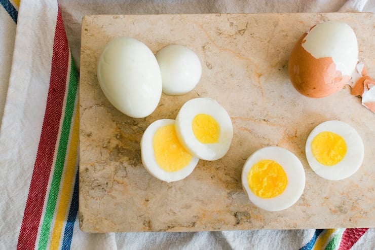 overhead shot of a marble cutting board with a partially peeled egg, a fully peeled egg, and a sliced hard-boiled egg. 