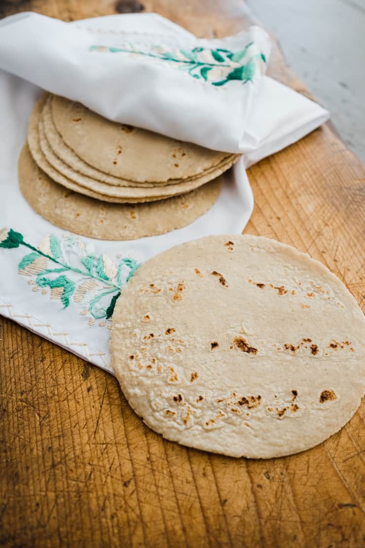 A stack of homemade corn tortillas on a brown wooden cutting board covered in a white embroidered Mexican linen