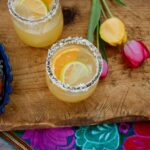 two skinny margaritas on a wooden cutting board with a colorful Mexican embroidered runner and yellow and pink tulips