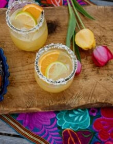 two skinny margaritas on a wooden cutting board with a colorful Mexican embroidered runner and yellow and pink tulips
