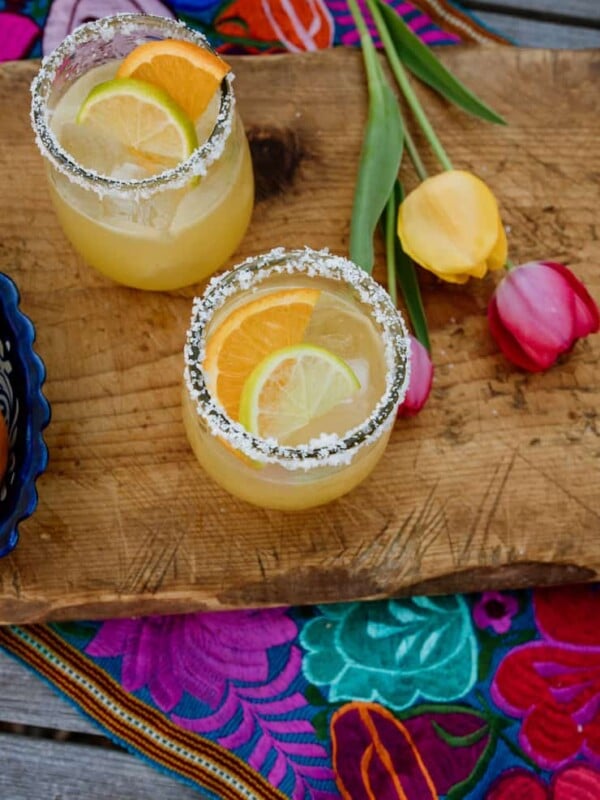 two skinny margaritas on a wooden cutting board with a colorful Mexican embroidered runner and yellow and pink tulips