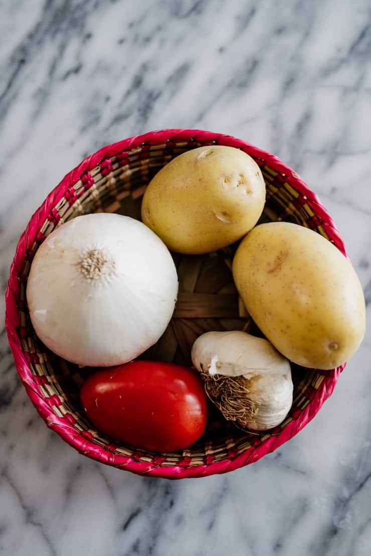 Mexican palm basket on a marble surface filled with a Roma tomato, onion, garlic and two potatoes 
