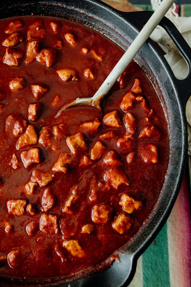 close up of red Chile pork stew in a cast iron pot with a wooden spoon 