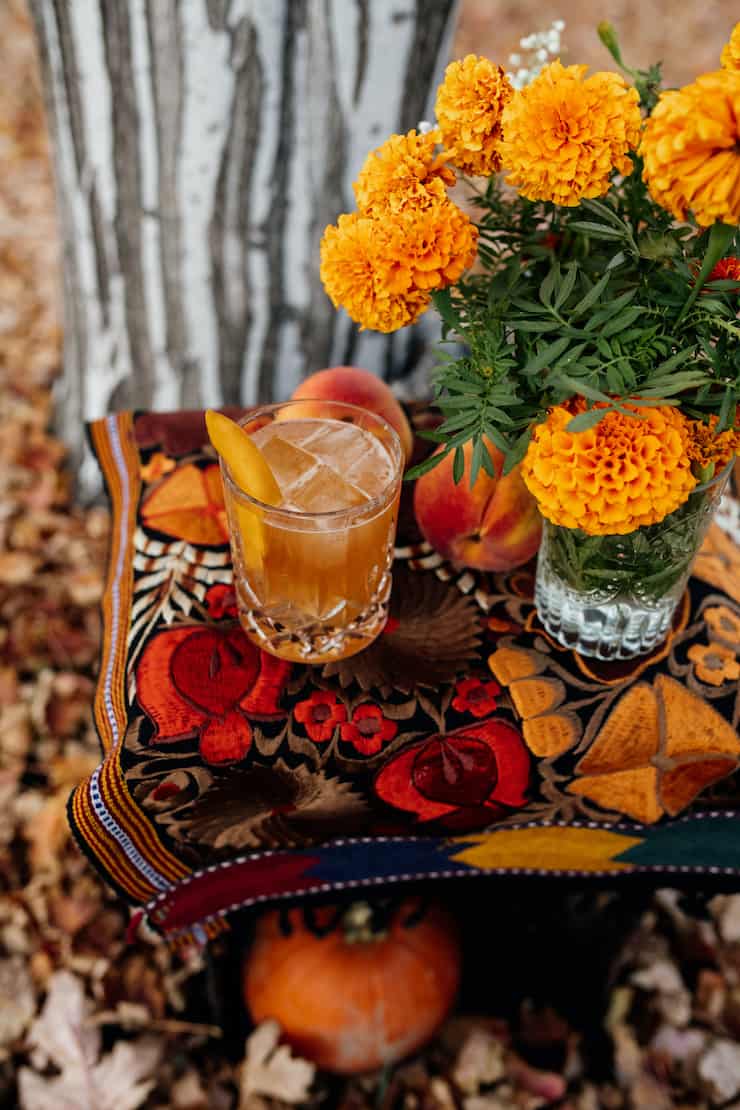 peach old fashioned cocktail in a cut crystal old fashioned glass on the rocks with a peach slice on a table with a bunch of marigolds, some fresh peaches and a striated grey and white tree trunk in the background