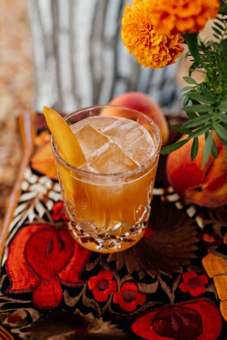 bourbon peach old fashioned with a peach garnish in a crystal glass on a red and orange tablescape with marigolds