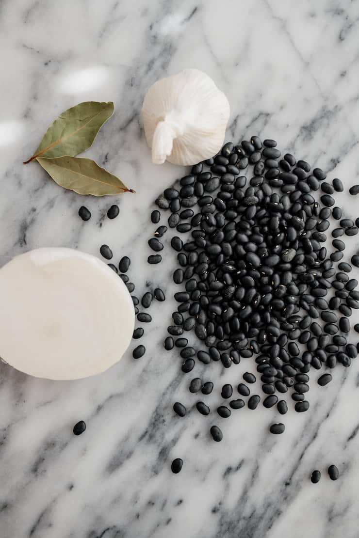 mise en place for homemade black beans - half an onion, 2 bay leaves, a head of garlic and dried black beans on a marble table