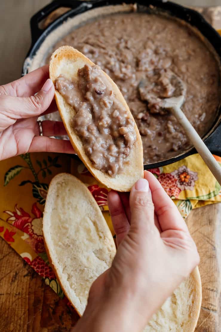 hands holding a toasted bolillo half with refried beans on top and a cast iron skillet with refried beans and a wood spoon in the background