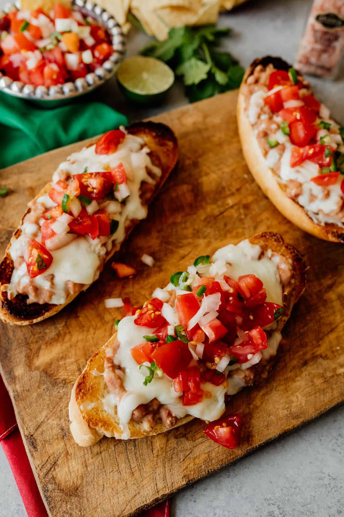 three molletes with refried beans, melted cheese and pico de gallo on a wooden cutting board.